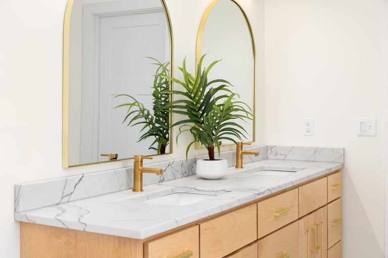 A modern double vanity bathroom featuring white marble countertops with gold faucets and arched mirrors hang above the sinks.