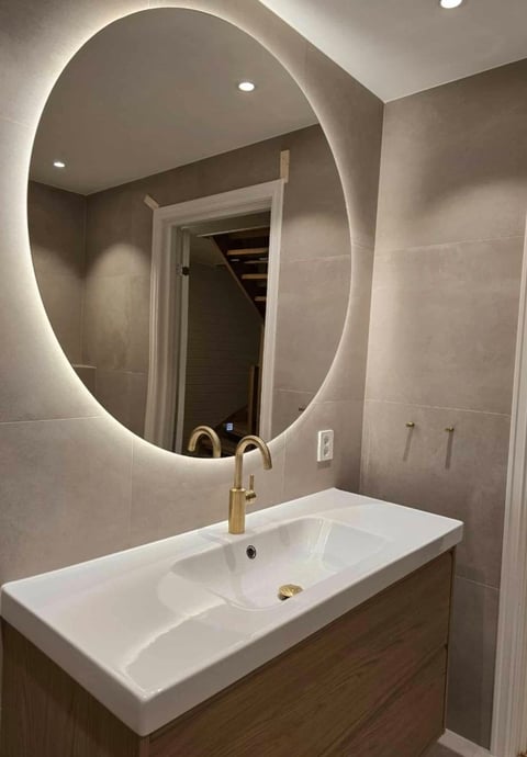Modern bathroom vanity with illuminated oval mirror, white rectangular sink, and brass gold faucet against taupe tile walls