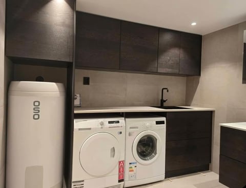 Modern laundry room with dark cabinets, white washer and dryer, and white water heater against beige tiled walls