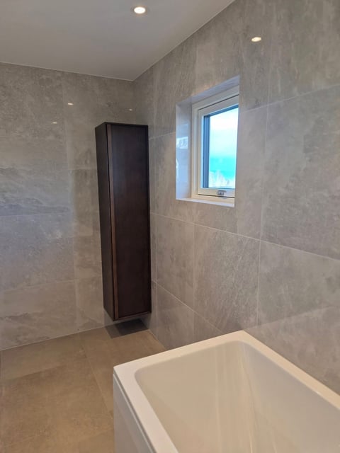 Modern bathroom interior with gray tile walls, white soaking tub, black tall cabinet, small window, and recessed ceiling lights