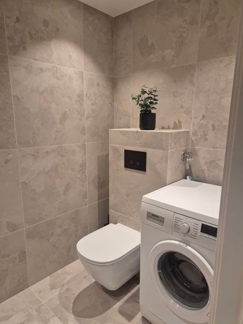 Modern bathroom with beige tile walls, white wall-mounted toilet, white washing machine beneath counter, and potted plant on shelf