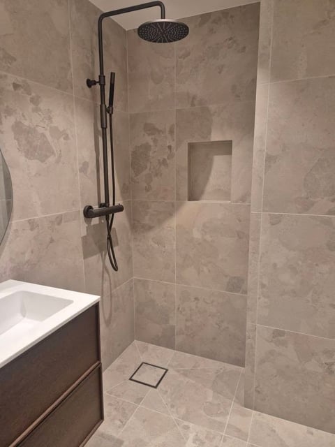 Modern bathroom shower with black rainfall showerhead and fixtures, beige tile walls, built-in niche, and white toilet visible
