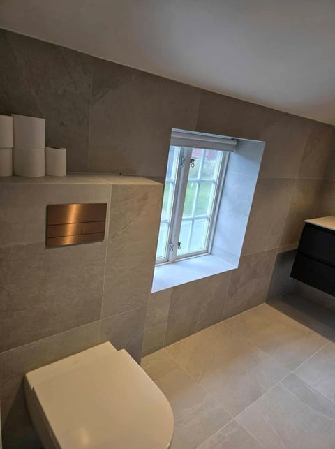 Modern bathroom with beige tile walls, white toilet, wall-mounted sink, and a window in the shower area