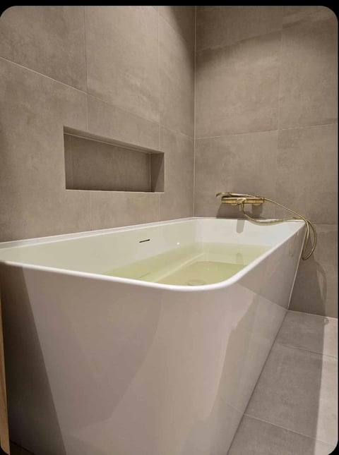 Modern white soaking tub filled with water in a beige tile bathroom with built-in niche and gold fixtures