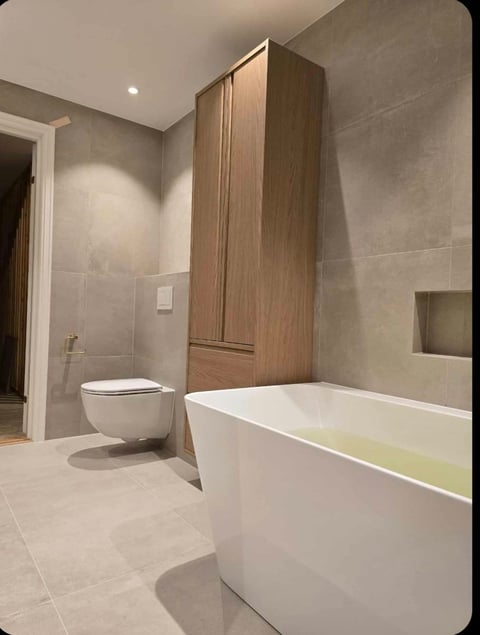 Modern bathroom with white freestanding bathtub, wall-mounted toilet, wooden cabinetry, and beige tile flooring and walls