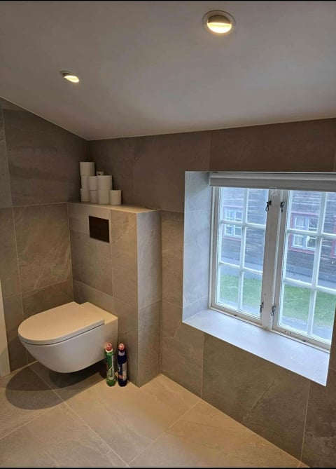 Modern bathroom interior with wall-mounted toilet, beige and gray tile, decorative items on shelf, and window with natural light