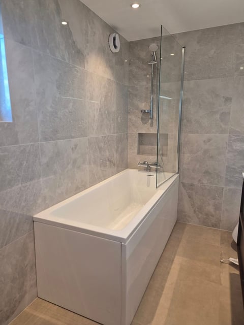 Modern bathroom with white rectangular bathtub and glass shower enclosure, gray tiled walls, and ceiling lights