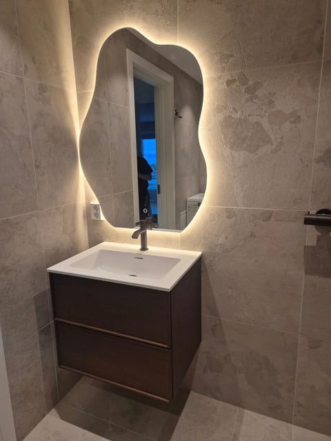 Modern bathroom vanity with backlit wavy mirror, dark wood cabinet, white square sink, and beige tile walls